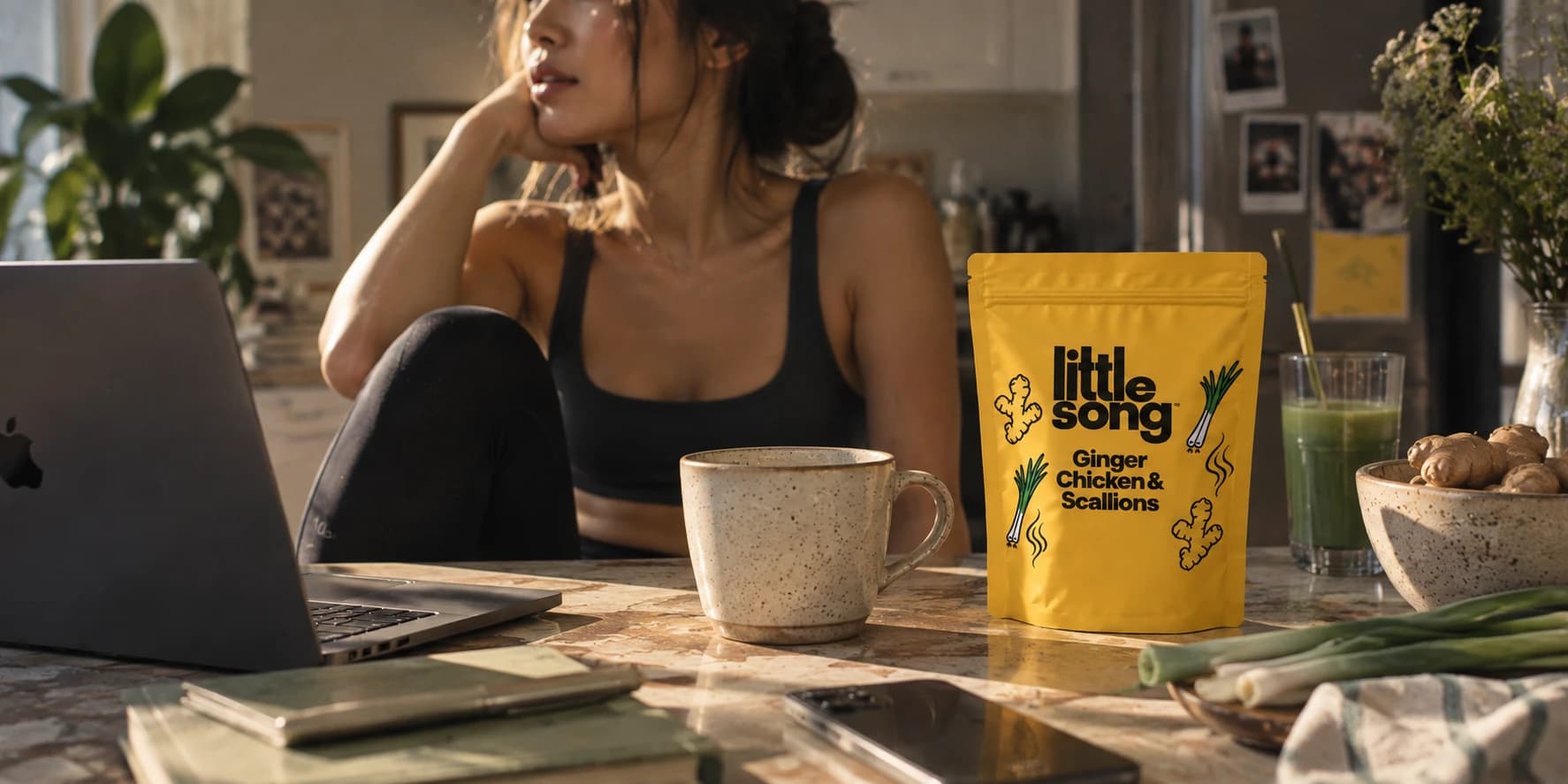 A woman takes a break at her sunlit kitchen table beside a mug and a pouch of Little Song Ginger Chicken & Scallions sipping broth